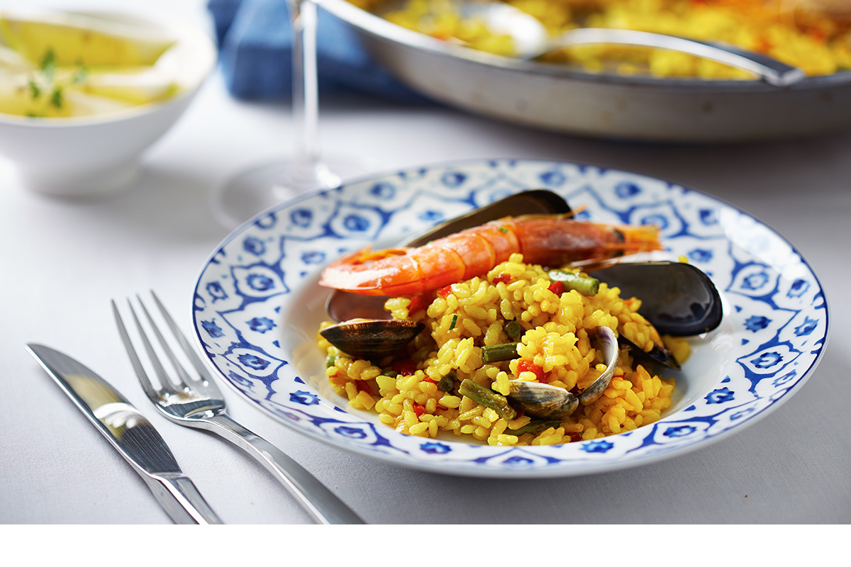 Ein Teller mit Meeresfrüchte-Paella, garniert mit einer Riesengarnele und Muscheln, serviert auf einem weiß-blau gemusterten Teller mit Besteck und Beilagen im Ristorante Mercato in Kloten.