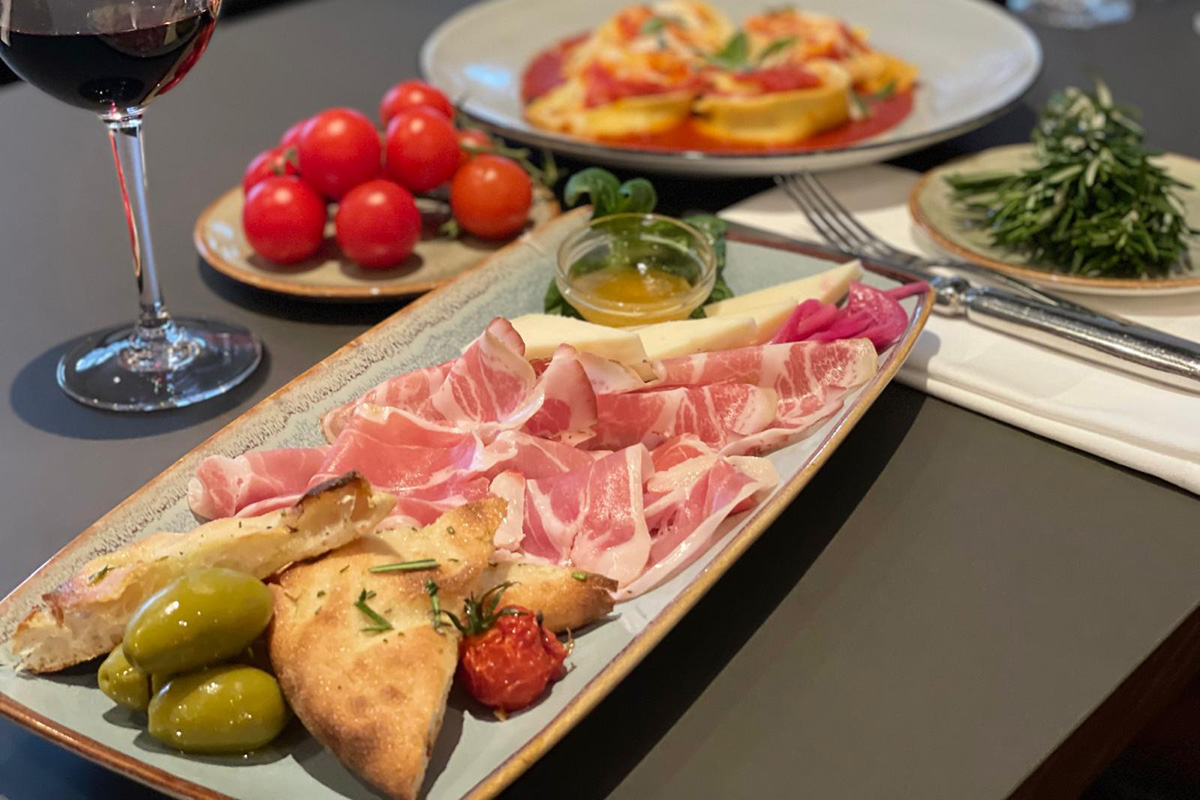 Platte mit Schinken und Käse mit einem Weinglas und Cherry-Tomaten im HIntergrund.