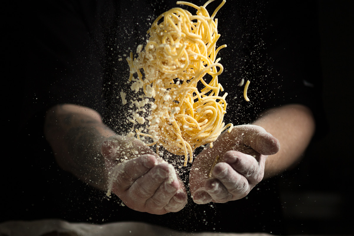 Mehlbedeckte Hände eines Kochs, die frische, dicke Spaghetti-Nudeln in die Luft werfen.